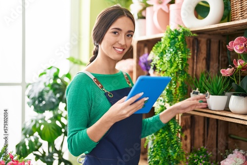 Young beautiful hispanic woman florist using touchpad holding plant of shelving at flower shop