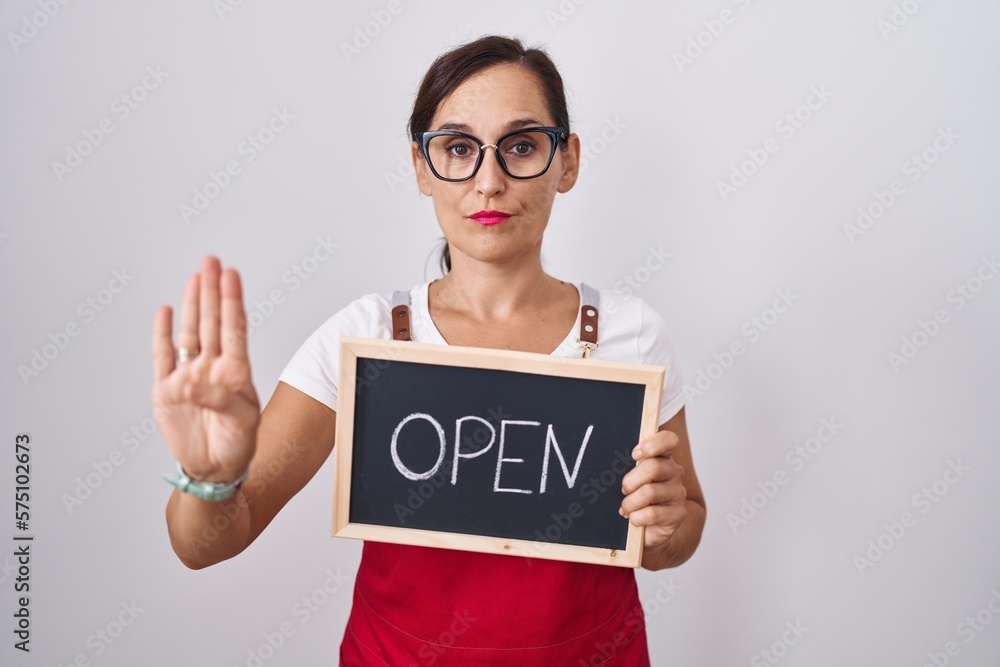 Middle age brunette woman holding banner with open text with open hand doing stop sign with serious and confident expression, defense gesture Middle age brunette woman holding banner with open text with open hand doing stop sign with serious and confident expression, defense gesture