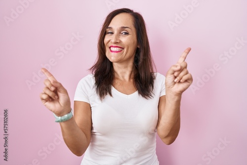 Middle age brunette woman standing over pink background smiling confident pointing with fingers to different directions. copy space for advertisement