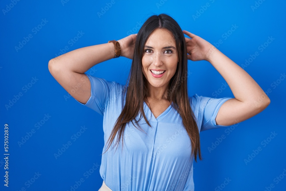 Young brunette woman standing over blue background relaxing and stretching, arms and hands behind head and neck smiling happy Young brunette woman standing over blue background relaxing and stretching, arms and hands behind head and neck smiling happy