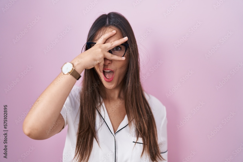 Young brunette woman wearing glasses standing over pink background peeking in shock covering face and eyes with hand, looking through fingers with embarrassed expression. Young brunette woman wearing glasses standing over pink background peeking in shock covering face and eyes with hand, looking through fingers with embarrassed expression.