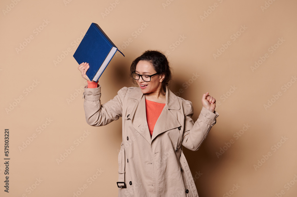 Cheerful joyful middle-aged woman, wearing beige coat and stylish spectacles, teacher educator celebrates the school year's end, dancing with a hardcover book in her hand, on isolated beige background Cheerful joyful middle-aged woman, wearing beige coat and stylish spectacles, teacher educator celebrates the school year's end, dancing with a hardcover book in her hand, on isolated beige background