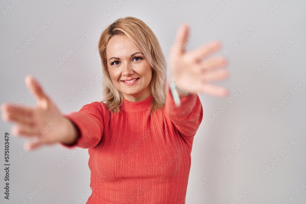 Blonde woman standing over isolated background looking at the camera smiling with open arms for hug. cheerful expression embracing happiness. Blonde woman standing over isolated background looking at the camera smiling with open arms for hug. cheerful expression embracing happiness.