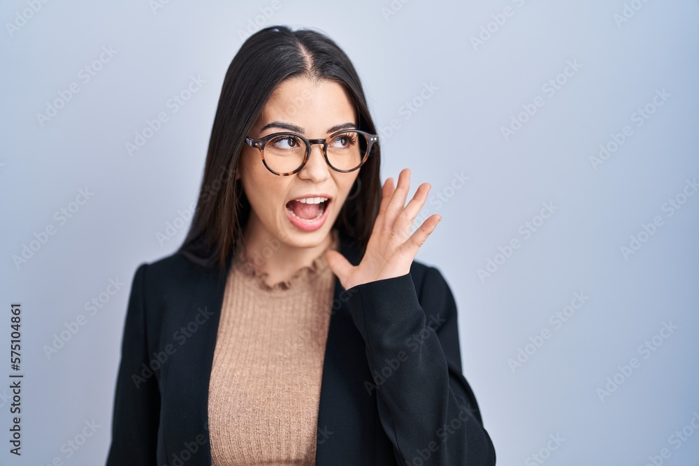 Young brunette woman standing over blue background shouting and screaming loud to side with hand on mouth. communication concept.