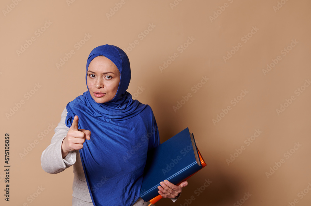 Confident strict Arab Muslim woman in blue hijab, pointing at camera, warning about the importance of reading book, standing with pile of books and looking at camera, isolated against beige background Confident strict Arab Muslim woman in blue hijab, pointing at camera, warning about the importance of reading book, standing with pile of books and looking at camera, isolated against beige background