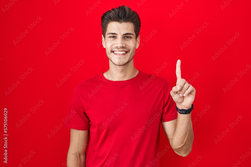 Young hispanic man standing over red background showing and pointing up with finger number one while smiling confident and happy. Young hispanic man standing over red background showing and pointing up with finger number one while smiling confident and happy.