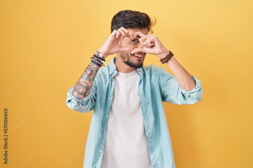 Young hispanic man with tattoos standing over yellow background doing heart shape with hand and fingers smiling looking through sign Young hispanic man with tattoos standing over yellow background doing heart shape with hand and fingers smiling looking through sign