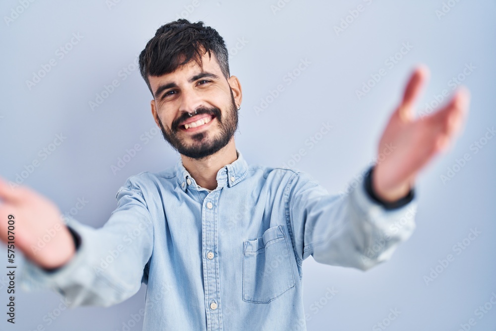 Young hispanic man with beard standing over blue background looking at the camera smiling with open arms for hug. cheerful expression embracing happiness. Young hispanic man with beard standing over blue background looking at the camera smiling with open arms for hug. cheerful expression embracing happiness.