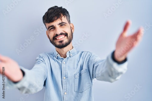 Young hispanic man with beard standing over blue background looking at the camera smiling with open arms for hug. cheerful expression embracing happiness. © Krakenimages.com