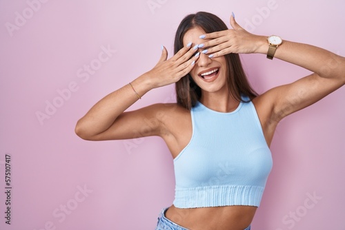 Young brunette woman standing over pink background covering eyes with hands smiling cheerful and funny. blind concept. © Krakenimages.com
