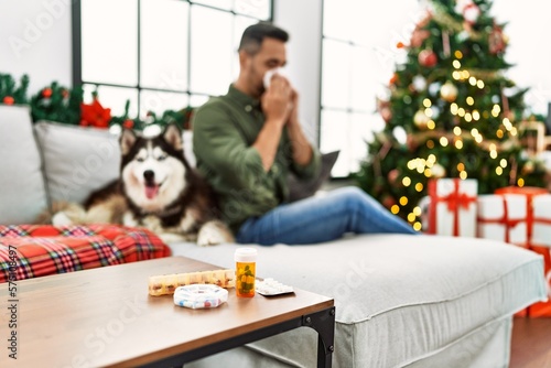Young hispanic man using tissue sitting on sofa with dog by christmas decor at home
