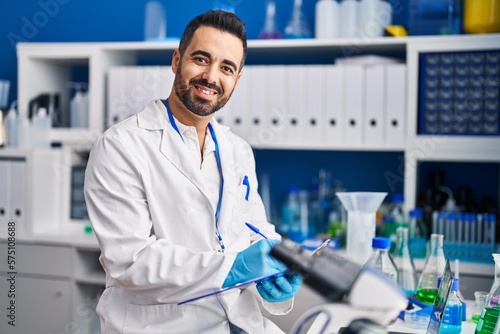 Young hispanic man scientist smiling confident writing on clipboard at laboratory
