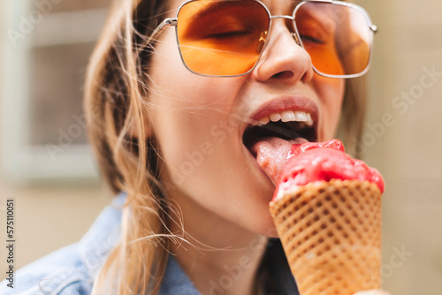 Close up happy young woman with delicious pink and white ice cream in waffle cone outdoors, closeup. Girl wear orange sunglasses in summer, spring or fall sunny day. Female lick ice cream and laughing
