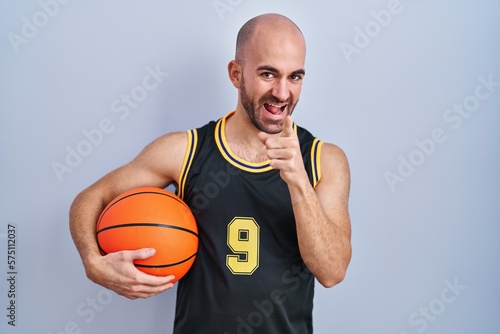 Young bald man with beard wearing basketball uniform holding ball pointing fingers to camera with happy and funny face. good energy and vibes.