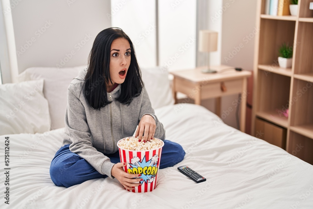 Hispanic woman eating popcorn watching a movie on the bed in shock face, looking skeptical and sarcastic, surprised with open mouth