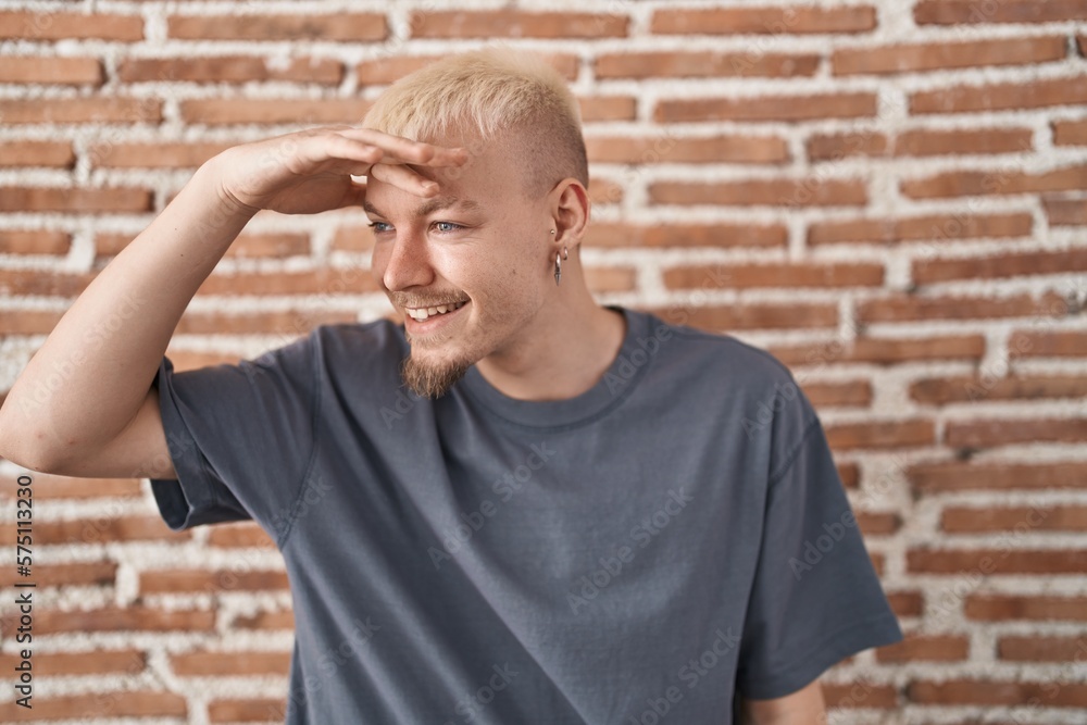 Young caucasian man standing over bricks wall very happy and smiling looking far away with hand over head. searching concept. Young caucasian man standing over bricks wall very happy and smiling looking far away with hand over head. searching concept.