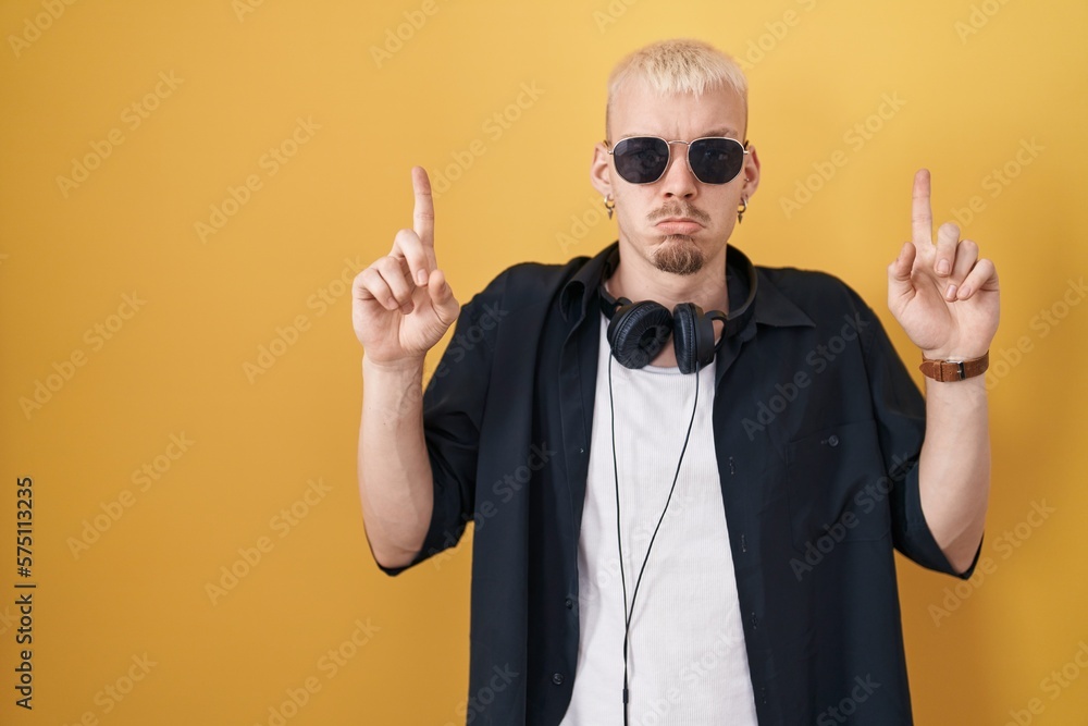 Young caucasian man wearing sunglasses standing over yellow background pointing up looking sad and upset, indicating direction with fingers, unhappy and depressed. Young caucasian man wearing sunglasses standing over yellow background pointing up looking sad and upset, indicating direction with fingers, unhappy and depressed.