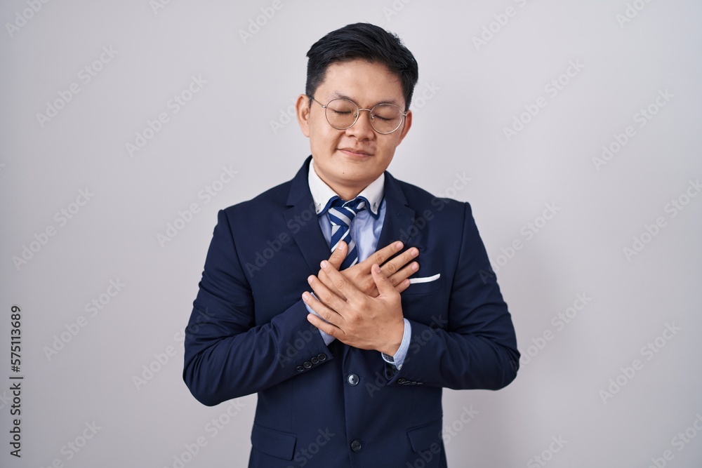 Young asian man wearing business suit and tie smiling with hands on chest with closed eyes and grateful gesture on face. health concept. Young asian man wearing business suit and tie smiling with hands on chest with closed eyes and grateful gesture on face. health concept.