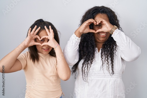 Mother and young daughter standing over white background doing heart shape with hand and fingers smiling looking through sign