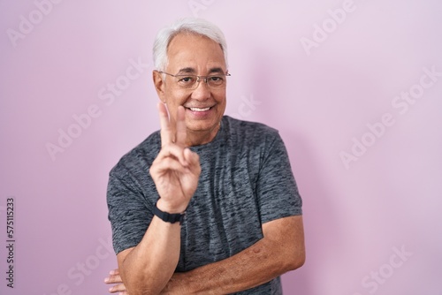 Middle age man with grey hair standing over pink background smiling with happy face winking at the camera doing victory sign with fingers. number two. © Krakenimages.com