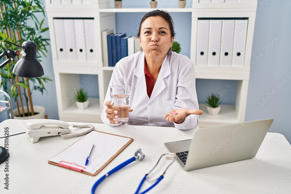 Hispanic mature doctor woman holding pill and glass of water puffing cheeks with funny face. mouth inflated with air, catching air. Hispanic mature doctor woman holding pill and glass of water puffing cheeks with funny face. mouth inflated with air, catching air.