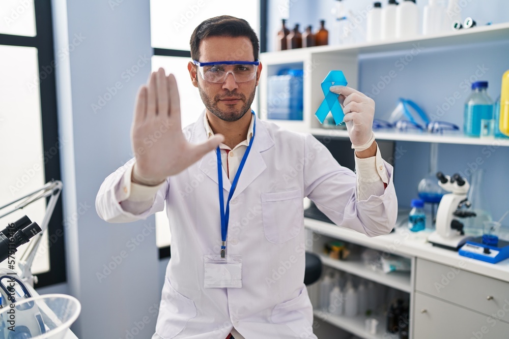 Young hispanic man with beard working at scientist laboratory holding blue ribbon with open hand doing stop sign with serious and confident expression, defense gesture Young hispanic man with beard working at scientist laboratory holding blue ribbon with open hand doing stop sign with serious and confident expression, defense gesture