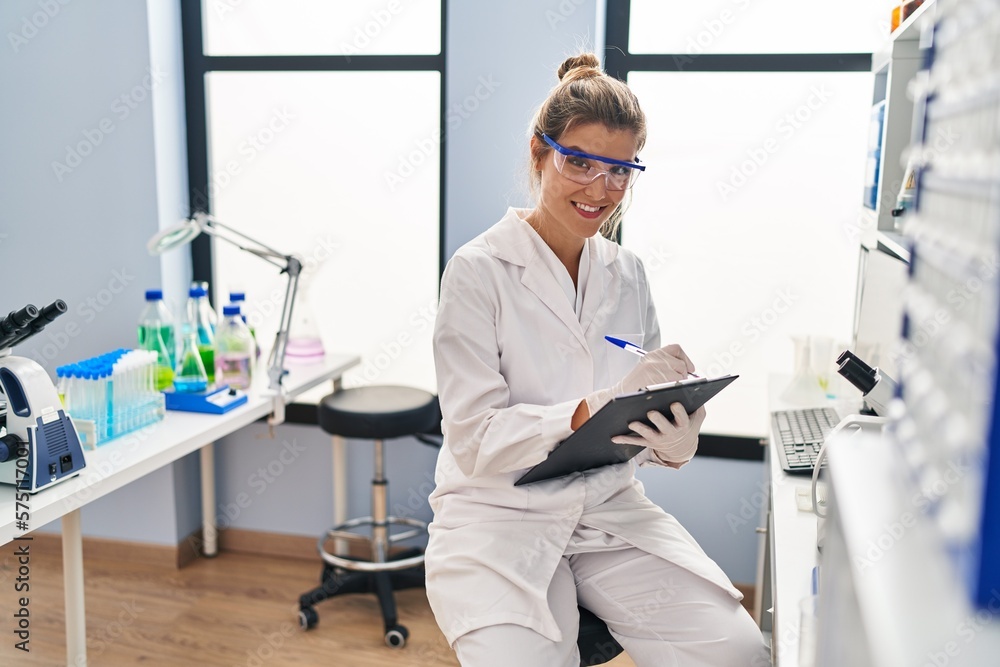 Young blonde woman wearing scientist uniform writing on clipboard at laboratory