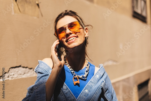 Beautiful young woman wearing denim shirt, blue top and orange sunglasses walking outdoors, talking on mobile phone. Portrait of stylish smiling woman ponytail calling on mobile phone on old street.