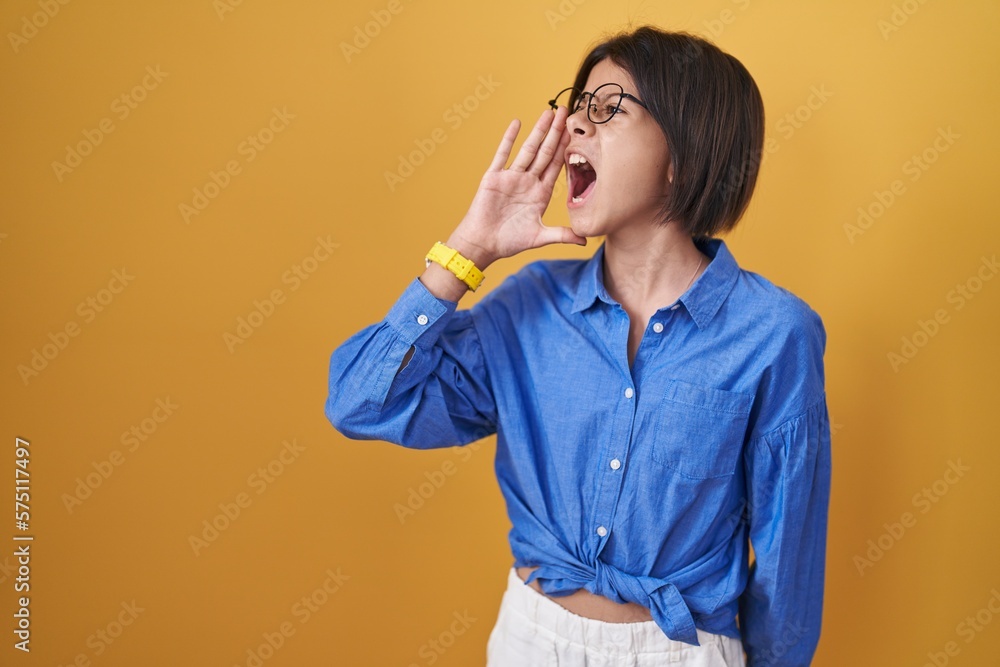 Young girl standing over yellow background shouting and screaming loud to side with hand on mouth. communication concept. Young girl standing over yellow background shouting and screaming loud to side with hand on mouth. communication concept.