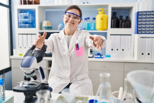Hispanic girl with down syndrome working at scientist laboratory looking at the camera smiling with open arms for hug. cheerful expression embracing happiness.