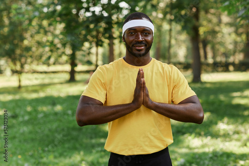 Waist up portrait of male yoga instructor looking at camera in park holding hands together by chest