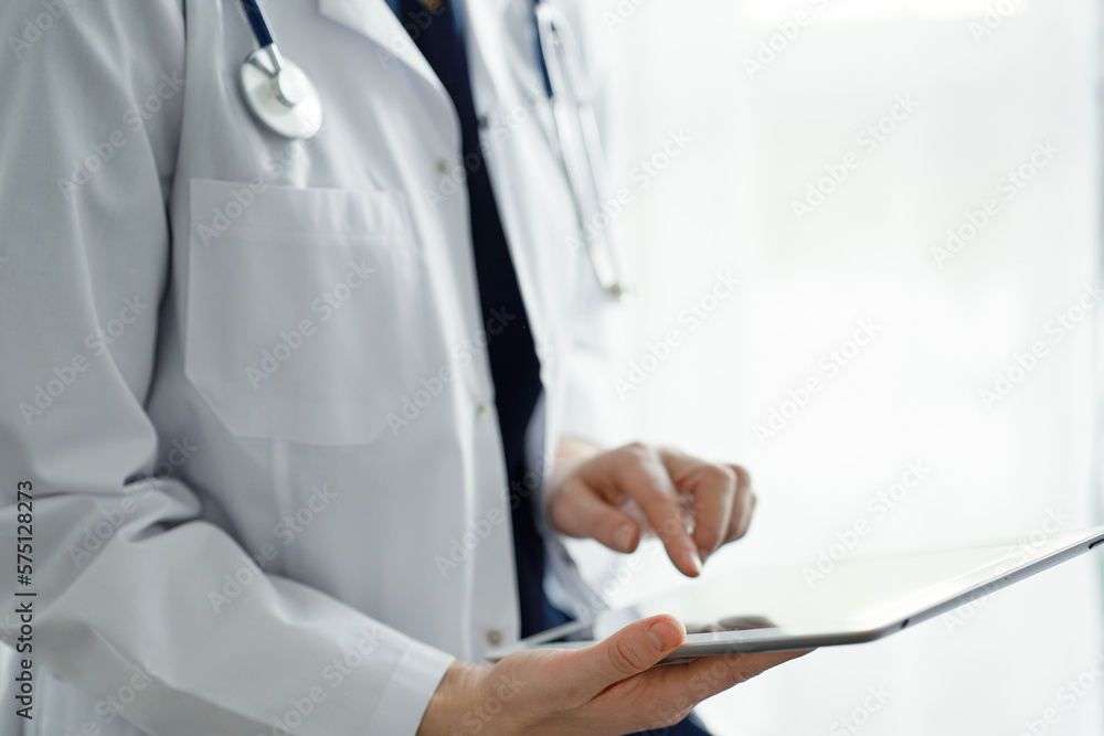 Doctor woman using tablet computer while standing near panorama window in clinic, close up. Physician or surgeon at work. Medicine concept Doctor woman using tablet computer while standing near panorama window in clinic, close up. Physician or surgeon at work. Medicine concept