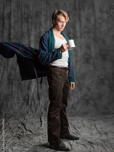 dude after a party in a housecoat with a shaggy head, Holding a mug in his hands and suffering from a hangover, in the studio on a gray background