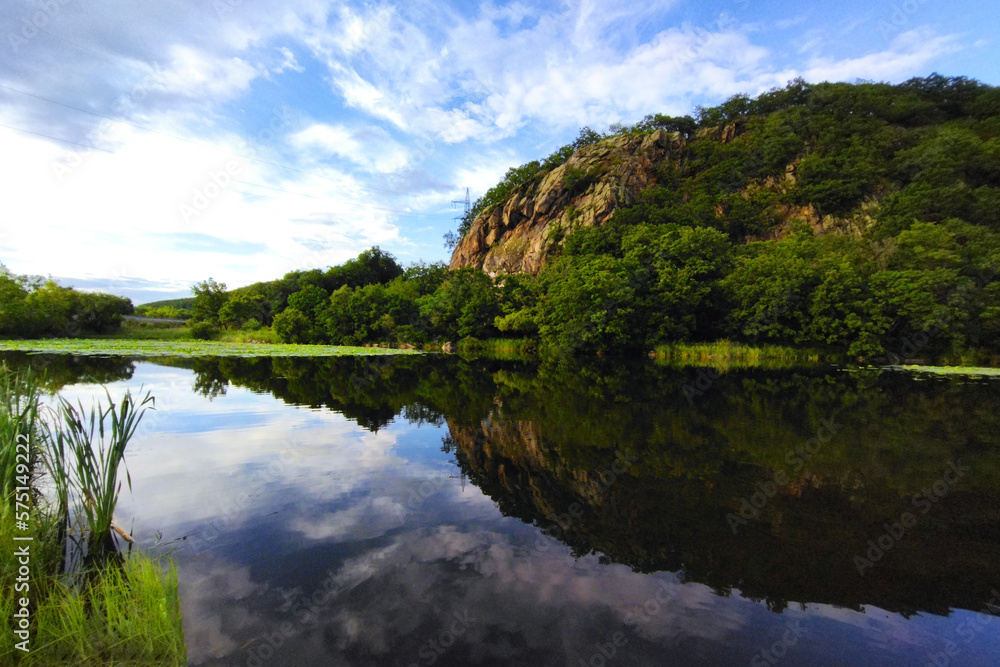 Lake and mountain range on a sunny summer day. The blue sky with clouds is reflected on the water surface. Beautiful summer landscape with a lake