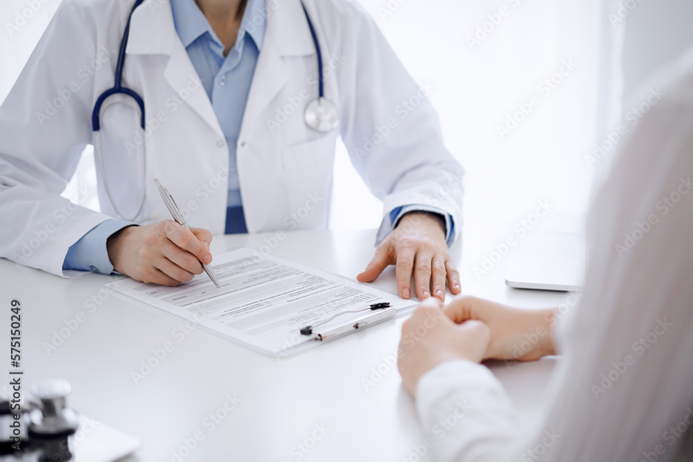 Doctor and patient sitting opposite each other at the desk in clinic. The focus is on female physician's hands filling up the medication history record form or checklist, close up. Medicine concept Doctor and patient sitting opposite each other at the desk in clinic. The focus is on female physician's hands filling up the medication history record form or checklist, close up. Medicine concept