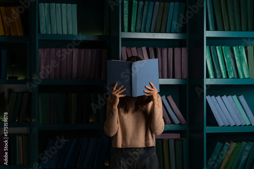A student in the library, preparing for exams, studying at the university. A young woman with a blue open book on the background of bookshelves.