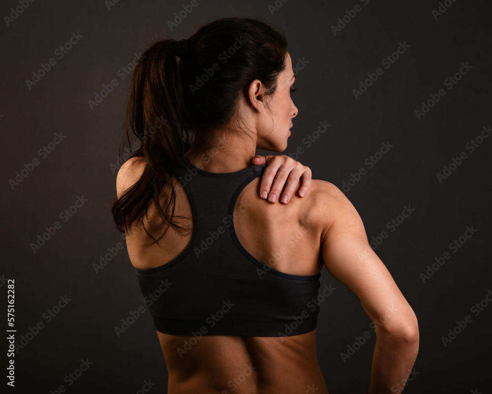 Fototapeta premium Serious female sporty muscular with ponytail doing stretching workout the shoulders, blades and arms in sport bra, holding the shoulder the hand on dark shadow studio colour empty space. Back view.