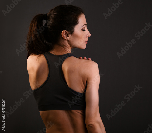Serious female sporty muscular with ponytail doing stretching workout the shoulders, blades and arms in sport bra, standing on dark shadow grey background with empty copy space. Back view. Lifestyle