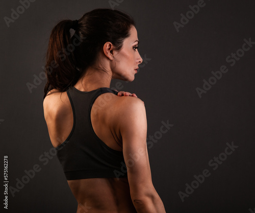 Serious female sporty muscular with ponytail doing stretching workout the shoulders, blades and arms in sport bra, standing on dark grey background with empty copy space. Back view. © nastia1983