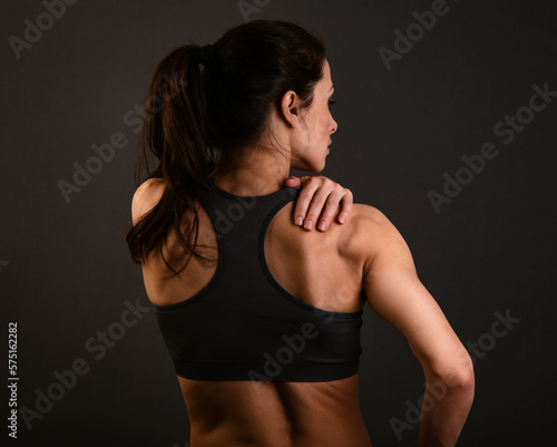 Serious female sporty muscular with ponytail doing stretching workout the shoulders, blades and arms in sport bra, holding the shoulder the hand on dark shadow studio colour empty space. Back view.