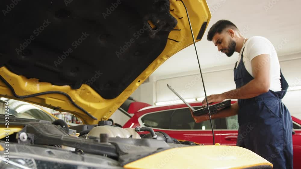 Young man car mechanic maintains vehicle using diagnostic laptop computer for checking engine problems, modern technology in the car repair shop. Service maintenance during engine repair Young man car mechanic maintains vehicle using diagnostic laptop computer for checking engine problems, modern technology in the car repair shop. Service maintenance during engine repair
