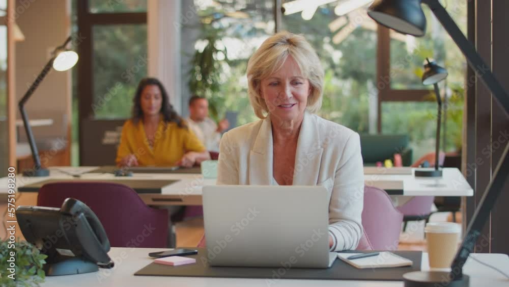 Mature businesswoman working on laptop at desk in modern office pauses to look out of the window and smile - shot in slow motion Mature businesswoman working on laptop at desk in modern office pauses to look out of the window and smile - shot in slow motion