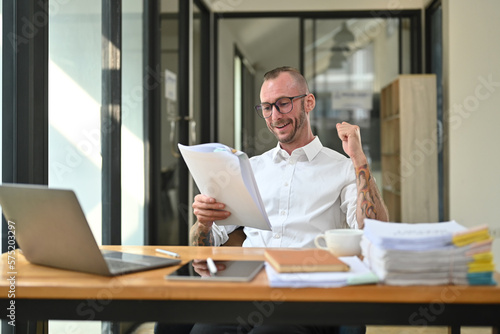 Happy businessman in a white shirt with tattooed celebrating success while reading documents in office
