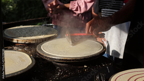 Making of crepes pancakes in street food center. A hand is making crepes outdoors on a metal griddle with wooden stick in outdoor