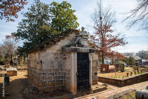 A statue of a man sitting in a chair with a hat in his lap on top of a mausoleum in a graveyard surrounded by lush green trees at the Oakland Cemetery with a blue sky and clouds in Atlanta Georgia USA photo