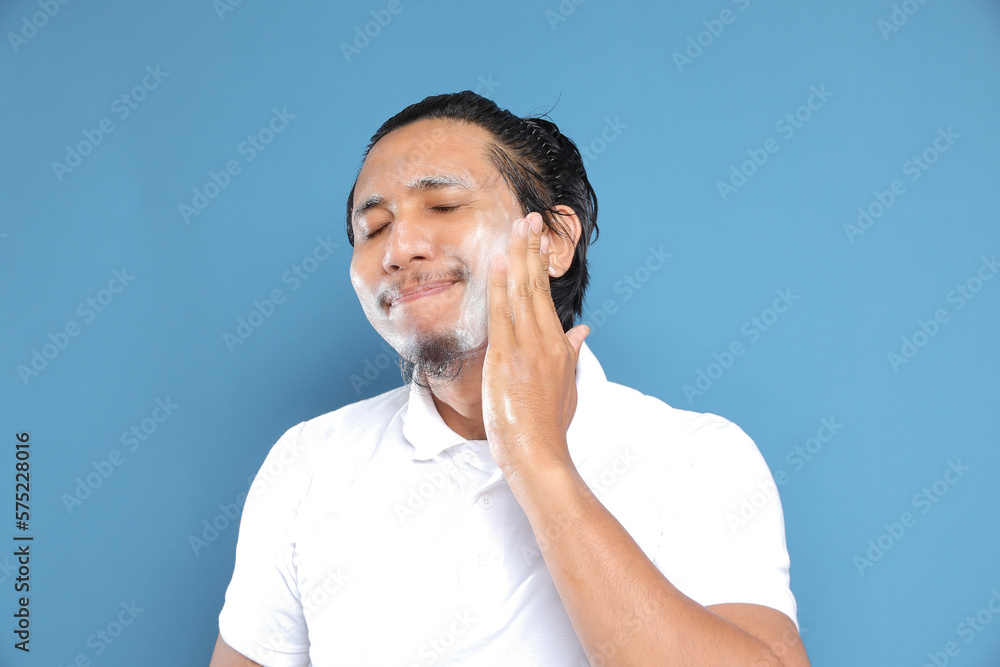 Close up portrait of young Asian man looking at camera, using facial clay mask, posing isolated over blue background. Beauty procedure and skincare routine concept. Horizontol shot Close up portrait of young Asian man looking at camera, using facial clay mask, posing isolated over blue background. Beauty procedure and skincare routine concept. Horizontol shot