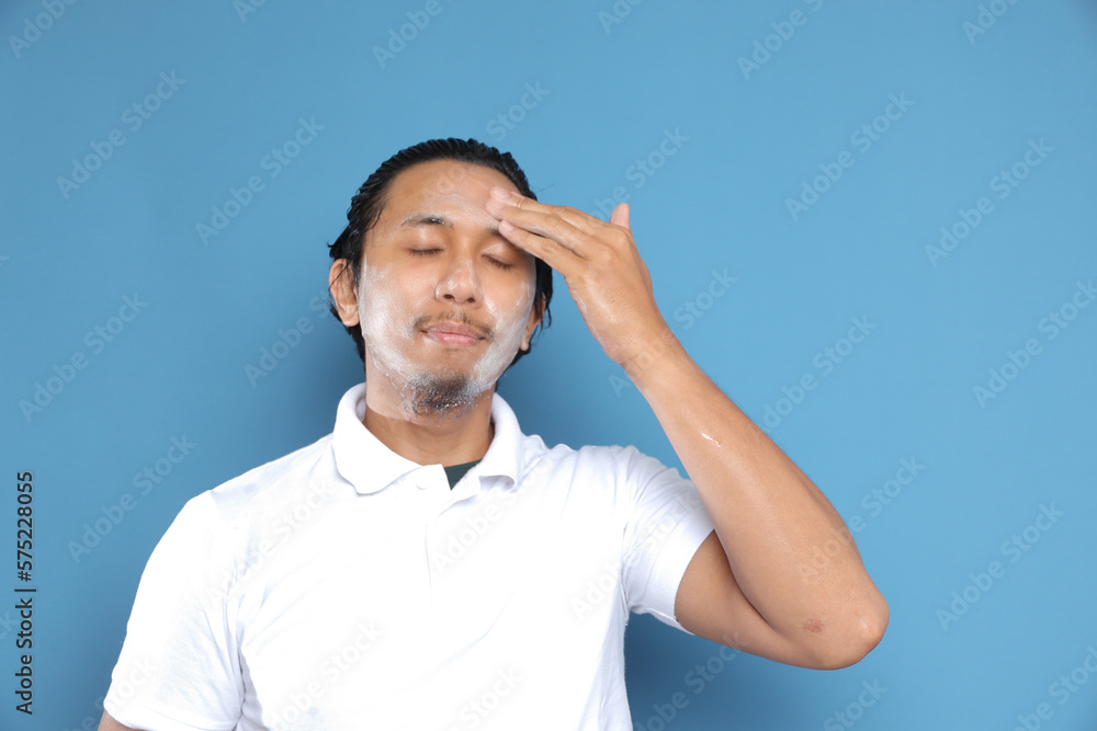 Fototapeta premium Close up portrait of young Asian man looking at camera, using facial clay mask, posing isolated over blue background. Beauty procedure and skincare routine concept. Horizontol shot