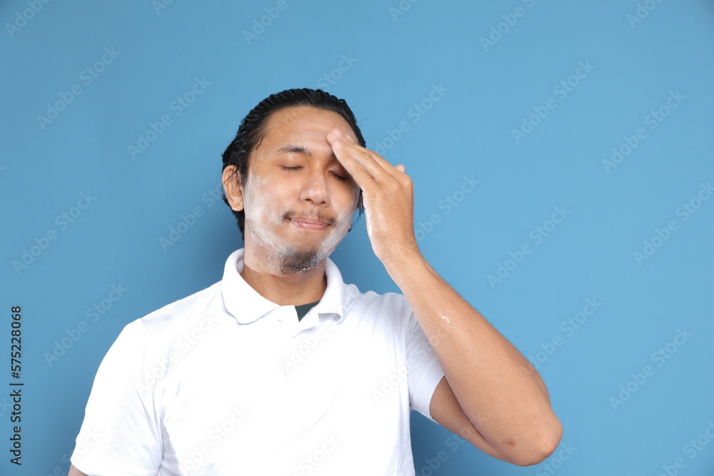 Close up portrait of young Asian man looking at camera, using facial clay mask, posing isolated over blue background. Beauty procedure and skincare routine concept. Horizontol shot Close up portrait of young Asian man looking at camera, using facial clay mask, posing isolated over blue background. Beauty procedure and skincare routine concept. Horizontol shot