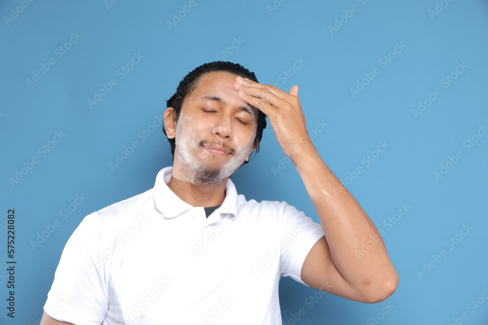 Naklejka premium Close up portrait of young Asian man looking at camera, using facial clay mask, posing isolated over blue background. Beauty procedure and skincare routine concept. Horizontol shot