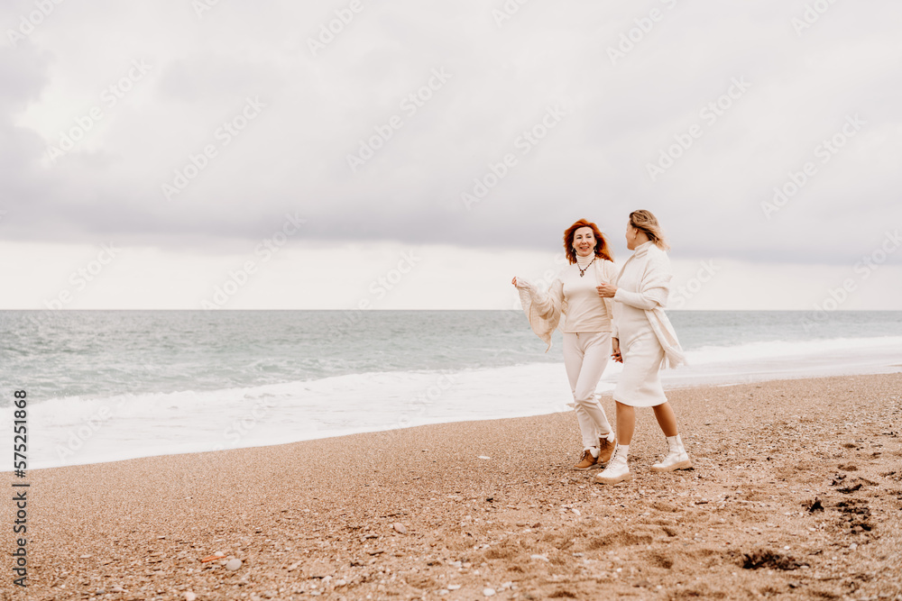 Women sea walk friendship spring. Two girlfriends, redhead and blonde, middle-aged walk along the sandy beach of the sea, dressed in white clothes. Against the backdrop of a cloudy sky and the winter Women sea walk friendship spring. Two girlfriends, redhead and blonde, middle-aged walk along the sandy beach of the sea, dressed in white clothes. Against the backdrop of a cloudy sky and the winter